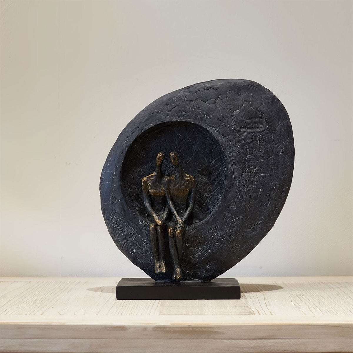 Abstract sculpture of two figures embracing on a heart-shaped platform against a neutral background