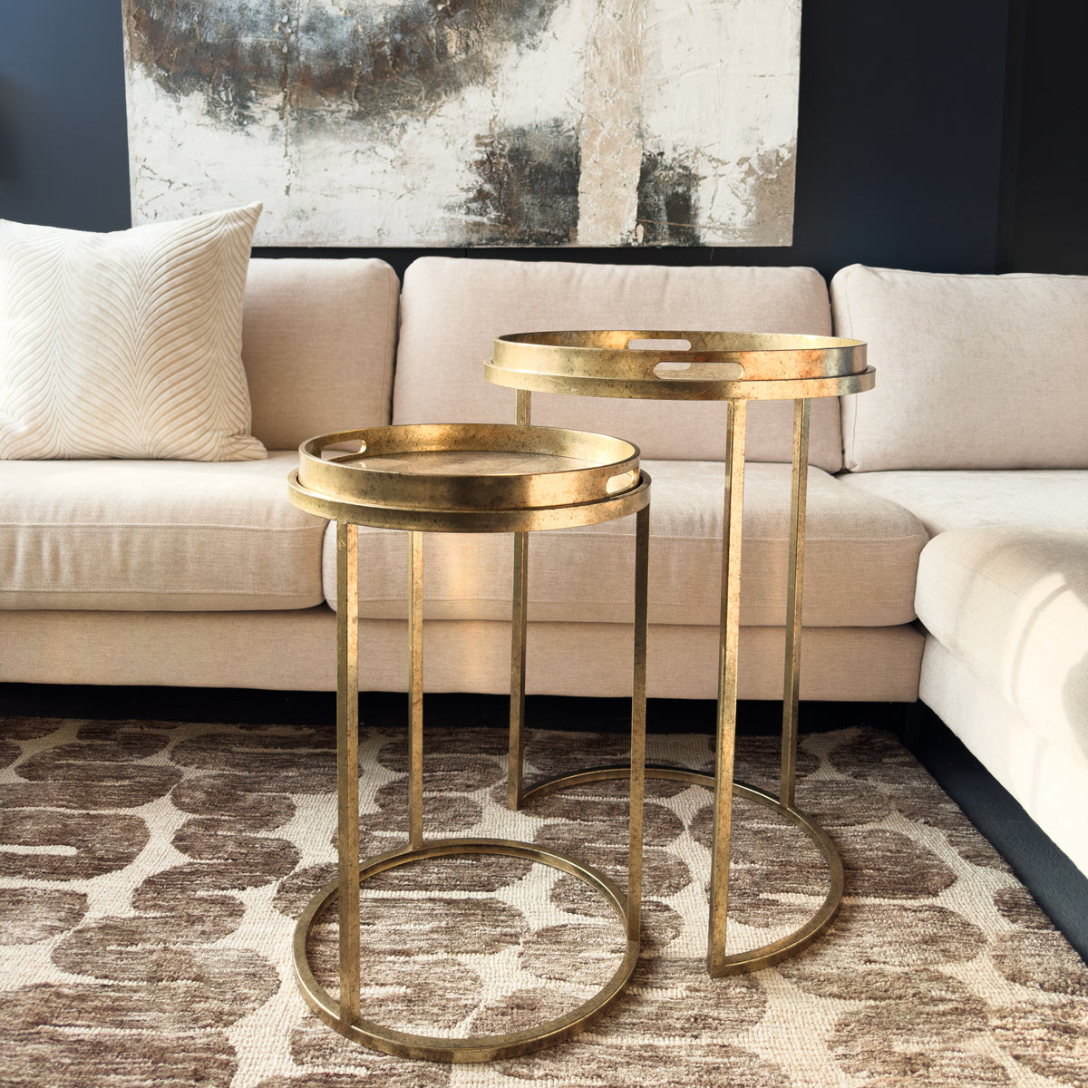 Two gold side tables in front of a beige sofa with a textured wall in the background.
