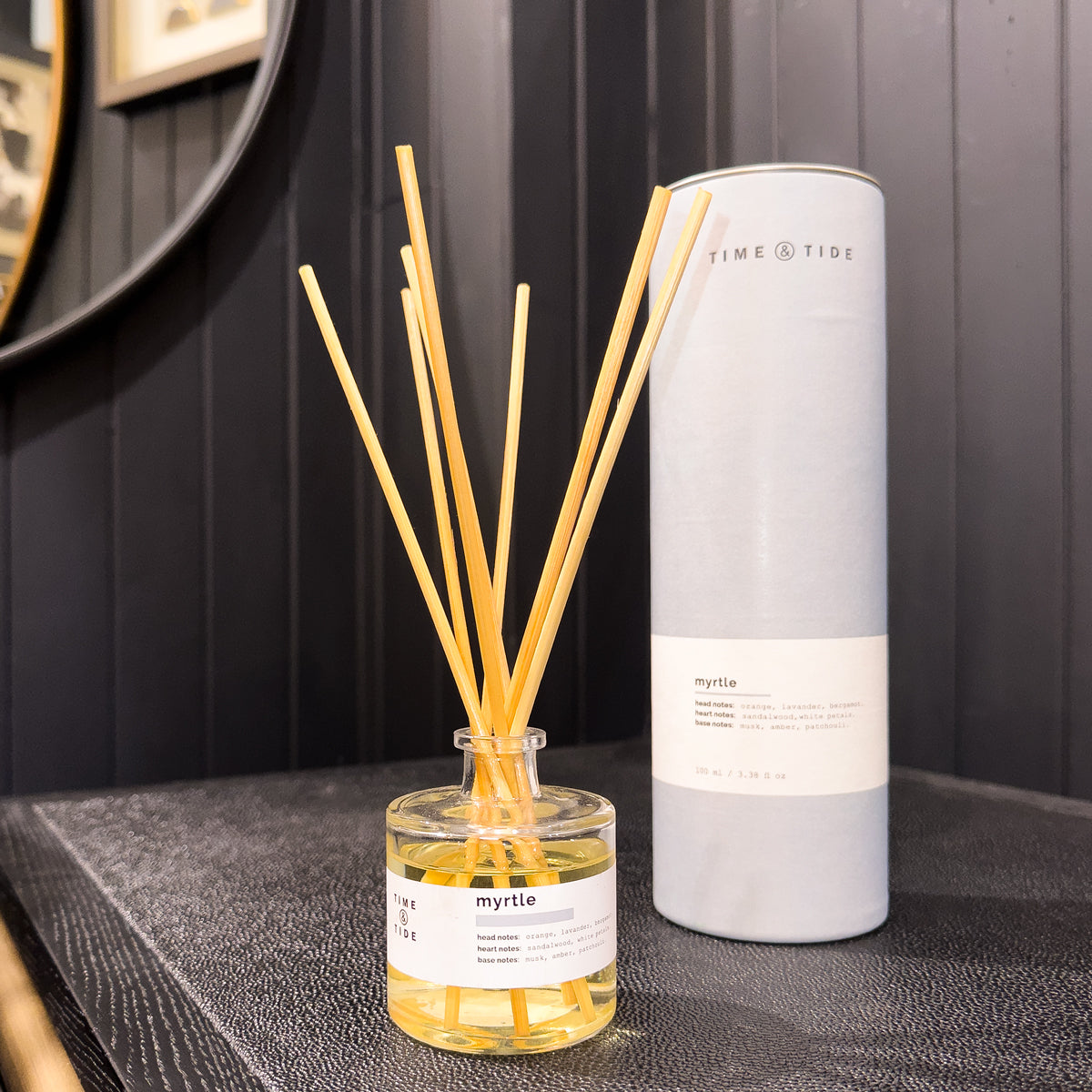 Diffuser with wooden sticks and a cylindrical container labeled 'Time & Tide' on a dark surface.