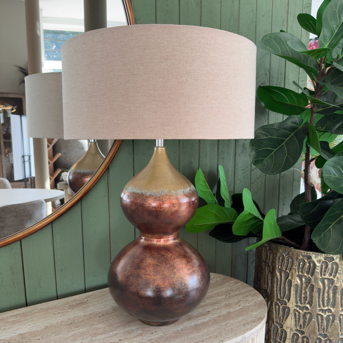 A warm copper and bronze table lamp with a wide beige shade sits on a pale stone console beside lush green plants, creating a calm, natural interior.