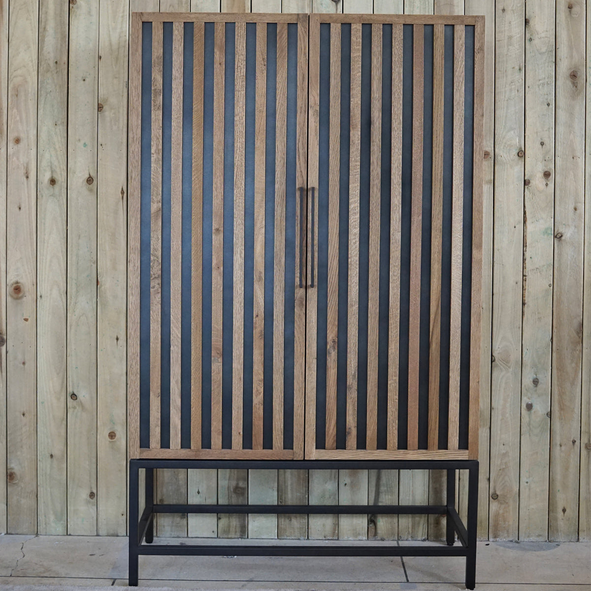 A tall slatted cabinet with oak wood slats and iron frame, featuring black inlay and minimalist handles.