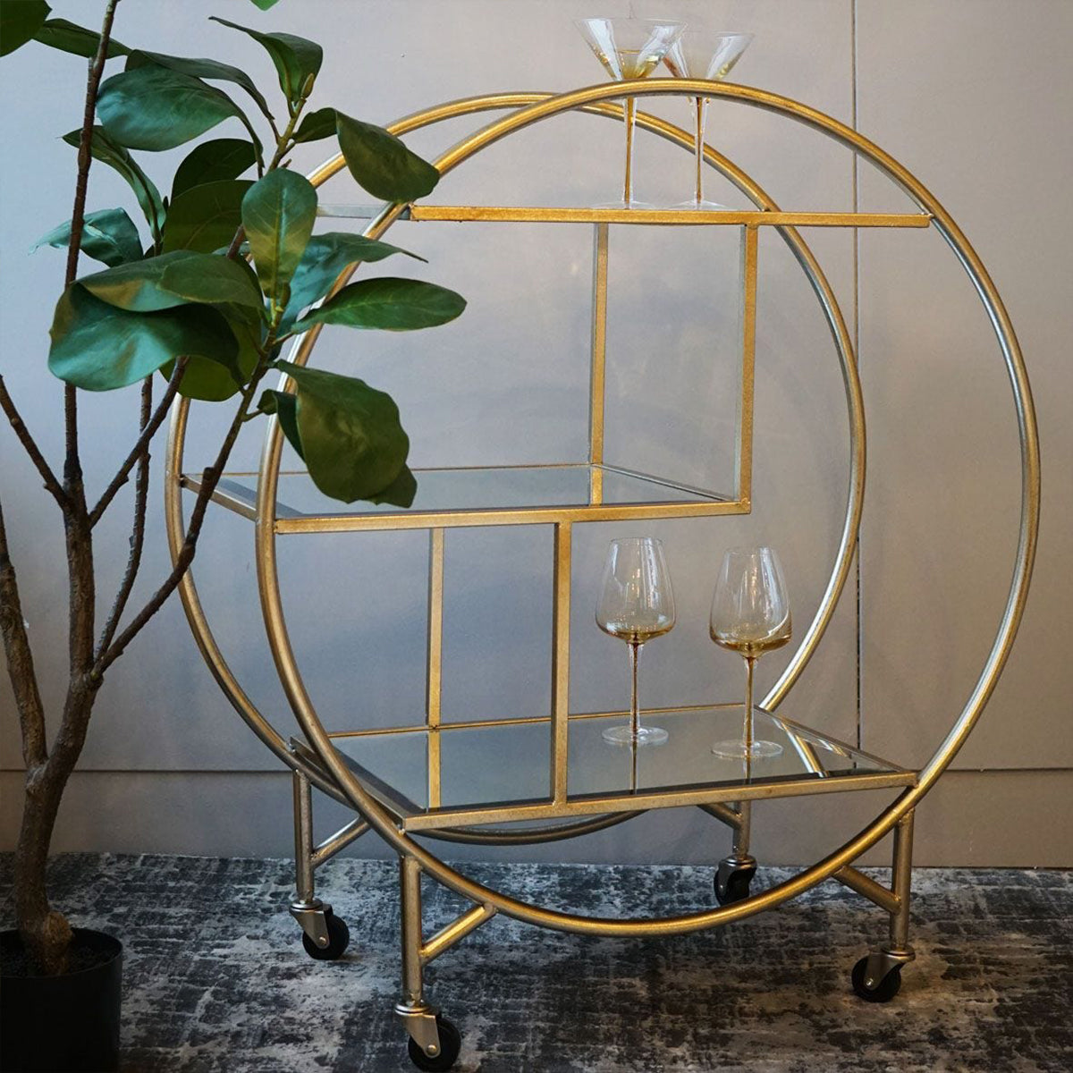 A champagne circular metal bar trolley with glass shelves and gold-colored metal frame, equipped with wheels, stored next to a potted plant.