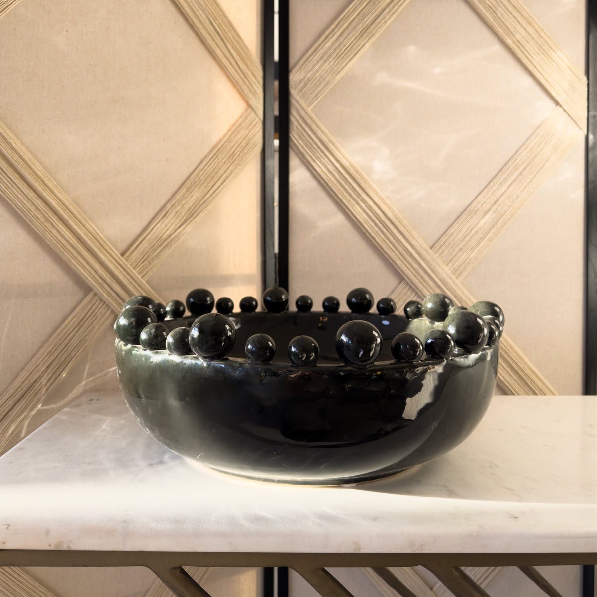 A charcoal black bowl with bobble details sitting on a white & gold marble console table, infront of a beige woven room divider.