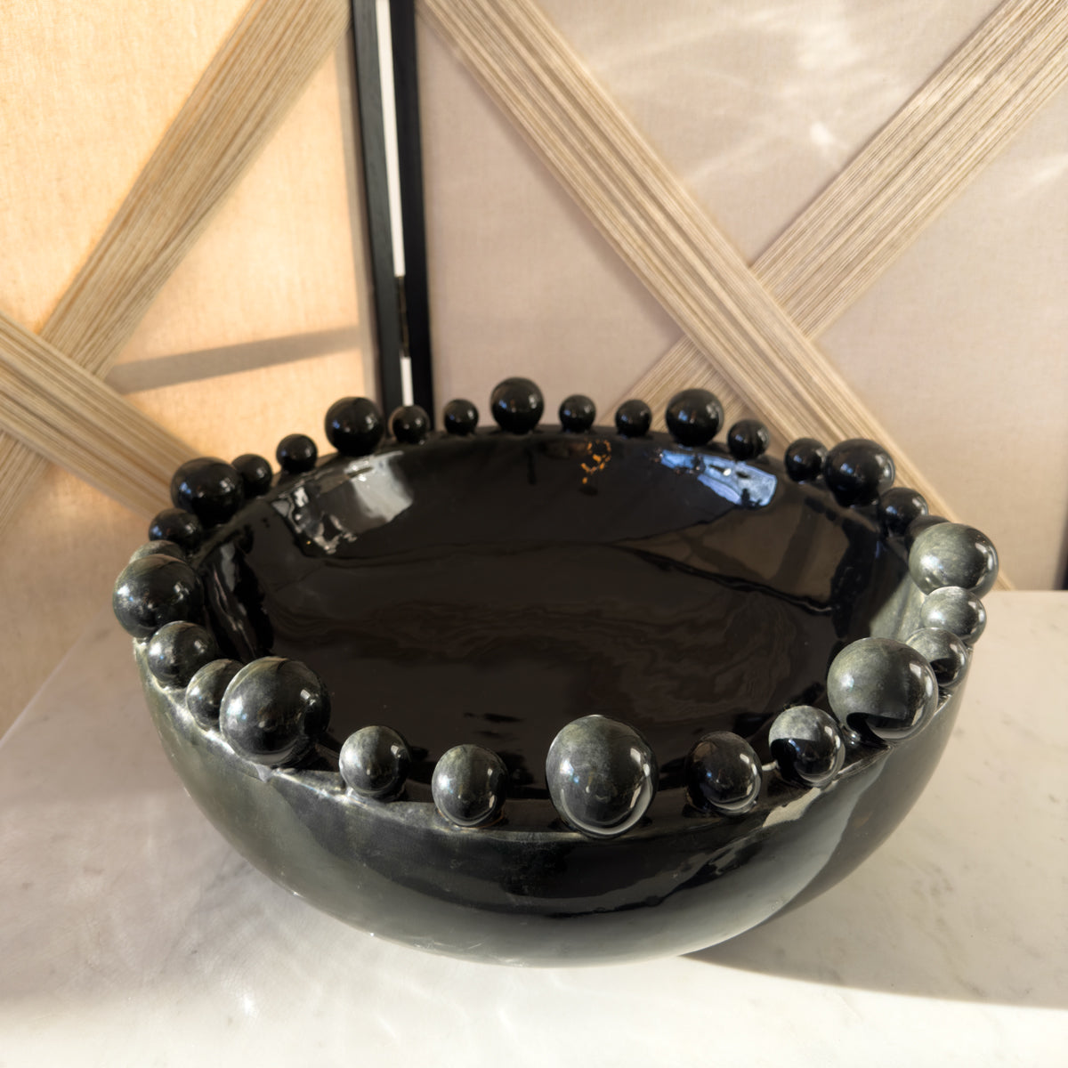A charcoal black bowl with bobble decorations around the rim, sitting on a white marble table infront of a woven room divider.