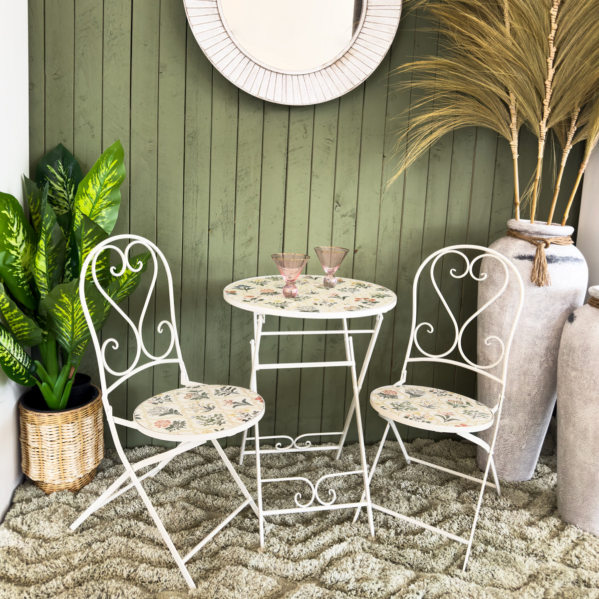 White metal bistro set with floral pattern against a green wooden wall.