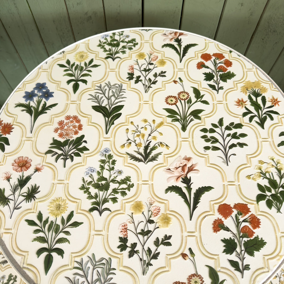 Round outdoor floral patterned table against a green wooden wall.