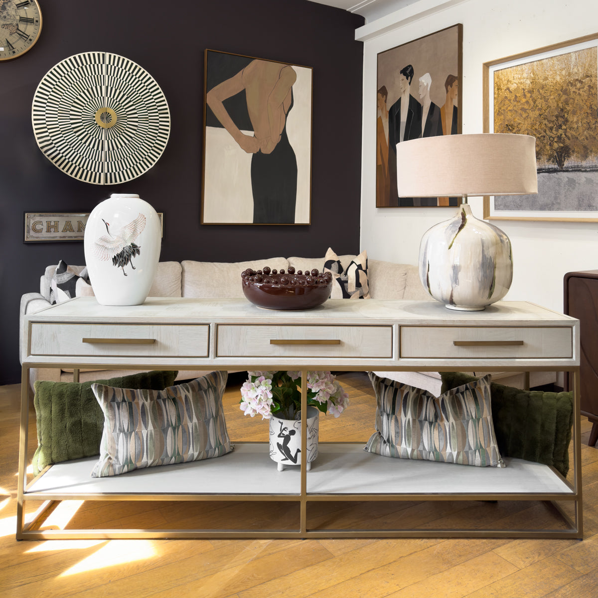 A modern console table in a styled living room setting, featuring a white parquet finish, three drawers with gold handles, and a lower shelf.