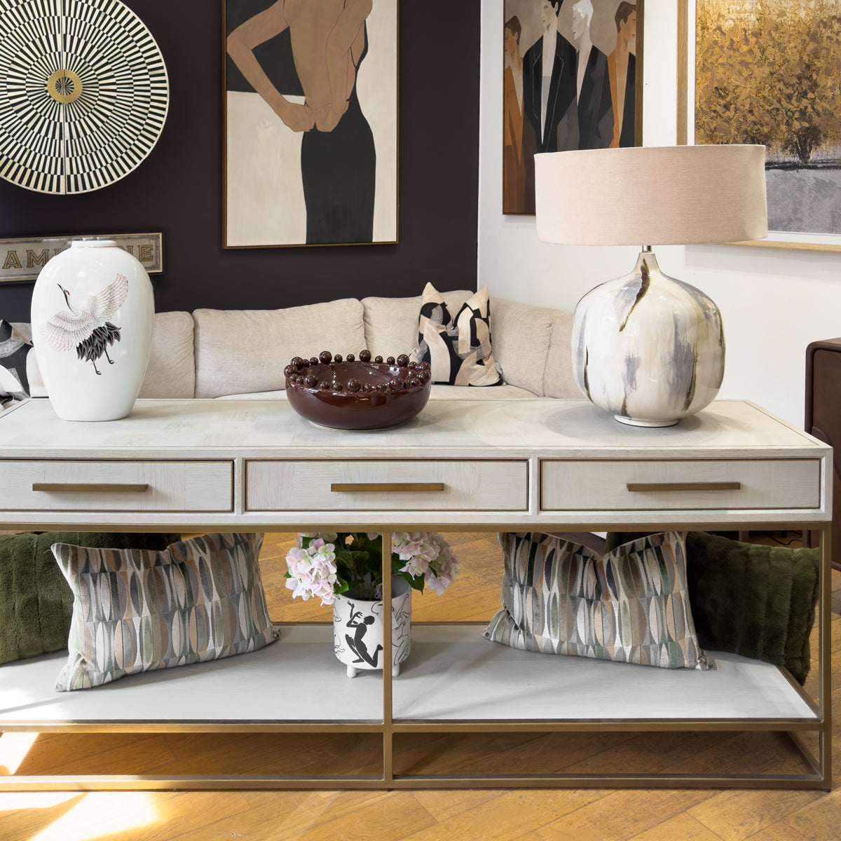 A modern console table in a styled living room setting. A lower shelf and a gold metal frame. The table is decorated with a white ceramic vase, a dark brown bowl, and a textured ceramic table lamp with a beige shade.