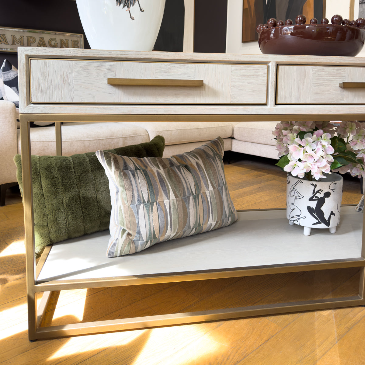 A three drawer console table with a light wood top, gold metal frame, and a white shelf, styled in a living room with decorative pillows and a vase of flowers.