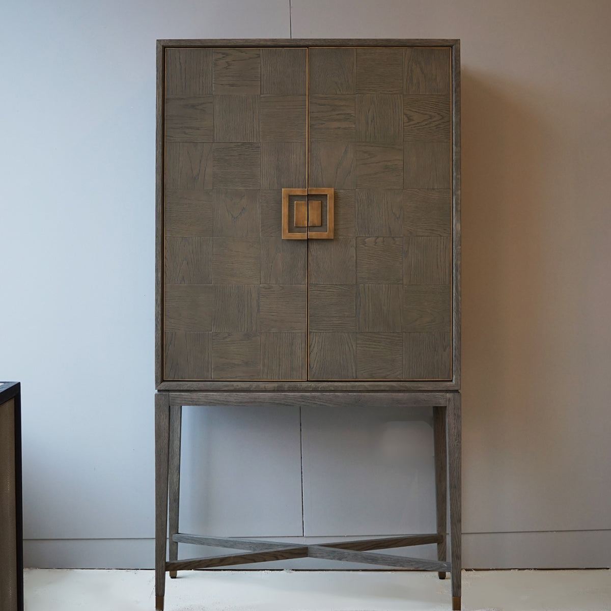 A greyed oak bar cabinet with brass inlay and handles, standing on a cross-braced frame, against a neutral background.