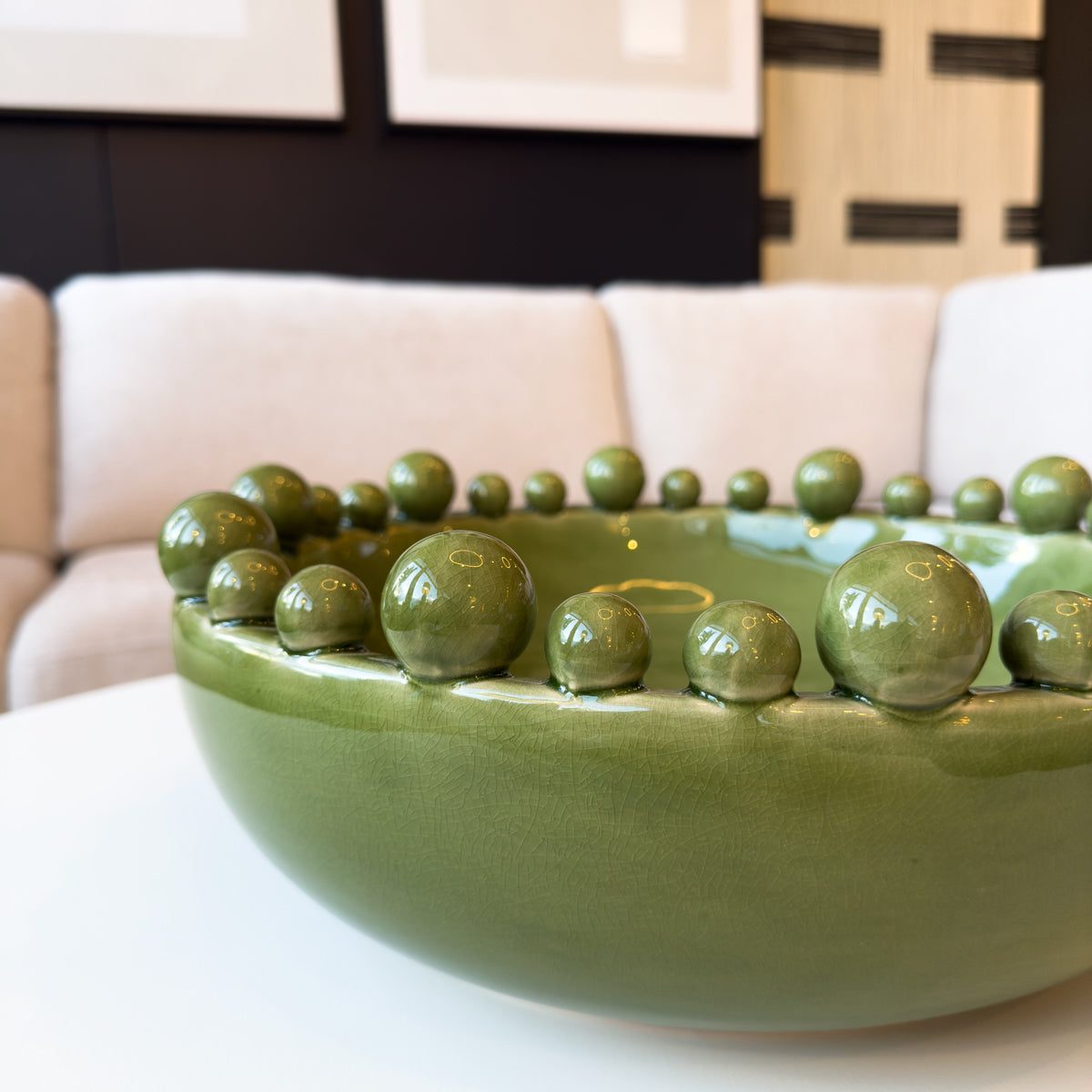 An olive green bobble surround bowl, sitting on a white coffee table infront of a neutral sofa.