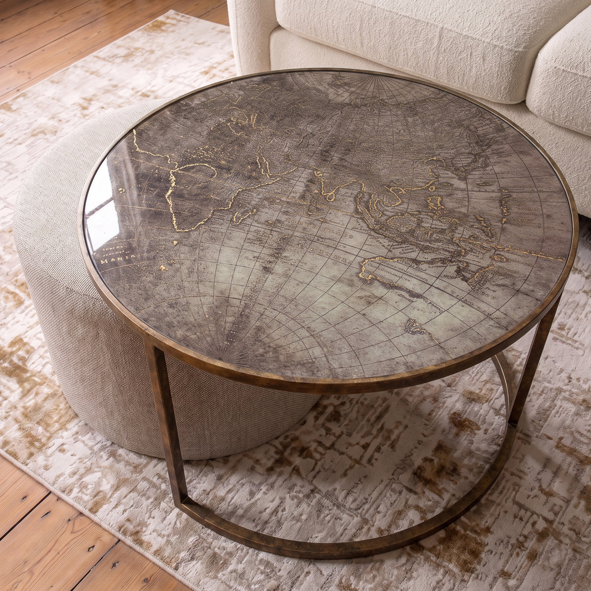 Circular coffee table with a vintage world map printed glass top and a curved gold metal base, styled in a living room next to a cream sofa and ottoman.