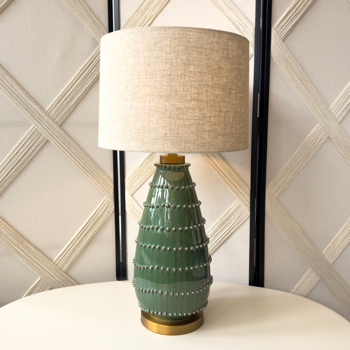 Green ceramic table lamp with beige lampshade against a decorative wall.