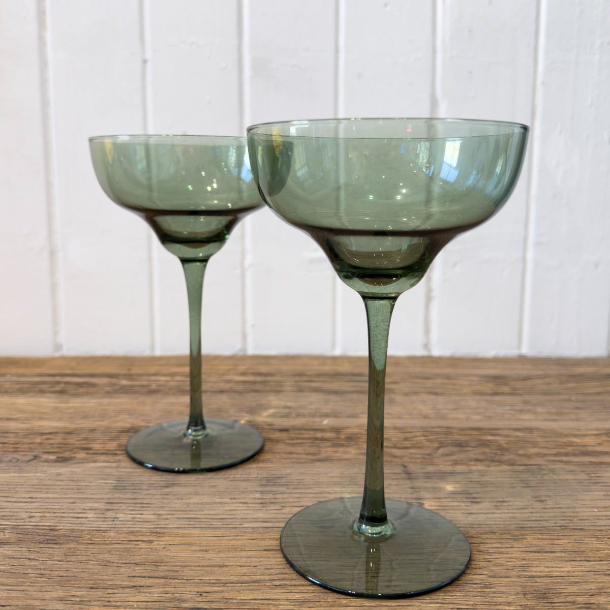 Two olive green martini glasses with a long stem, displayed on a wooden surface against a white background.