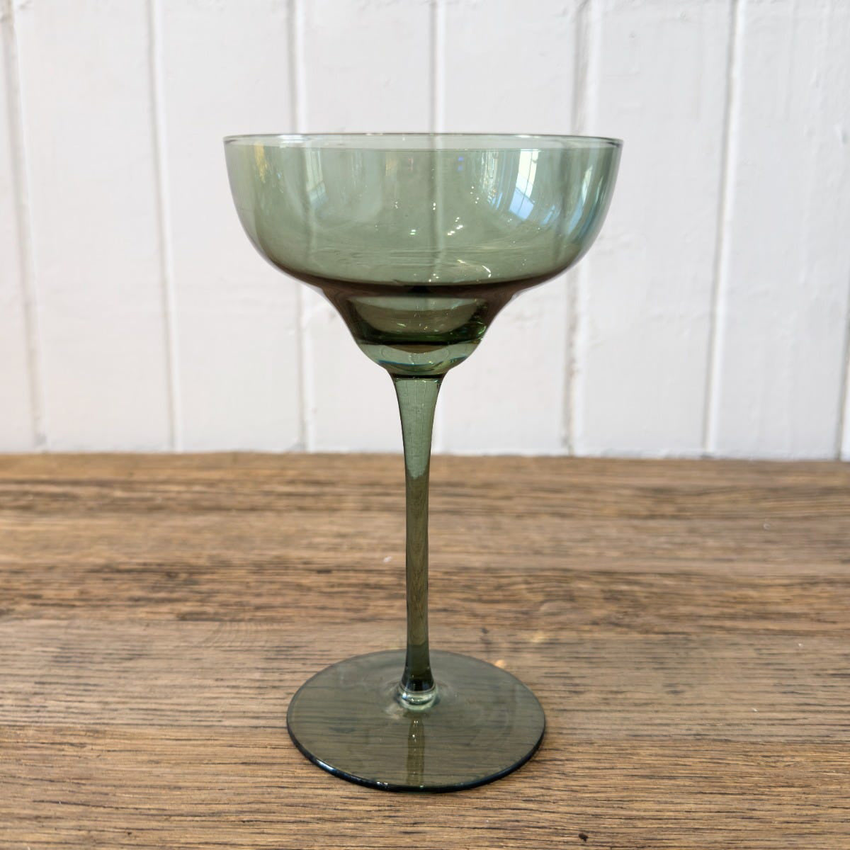 Green glass coupe on a wooden surface with a white paneled background