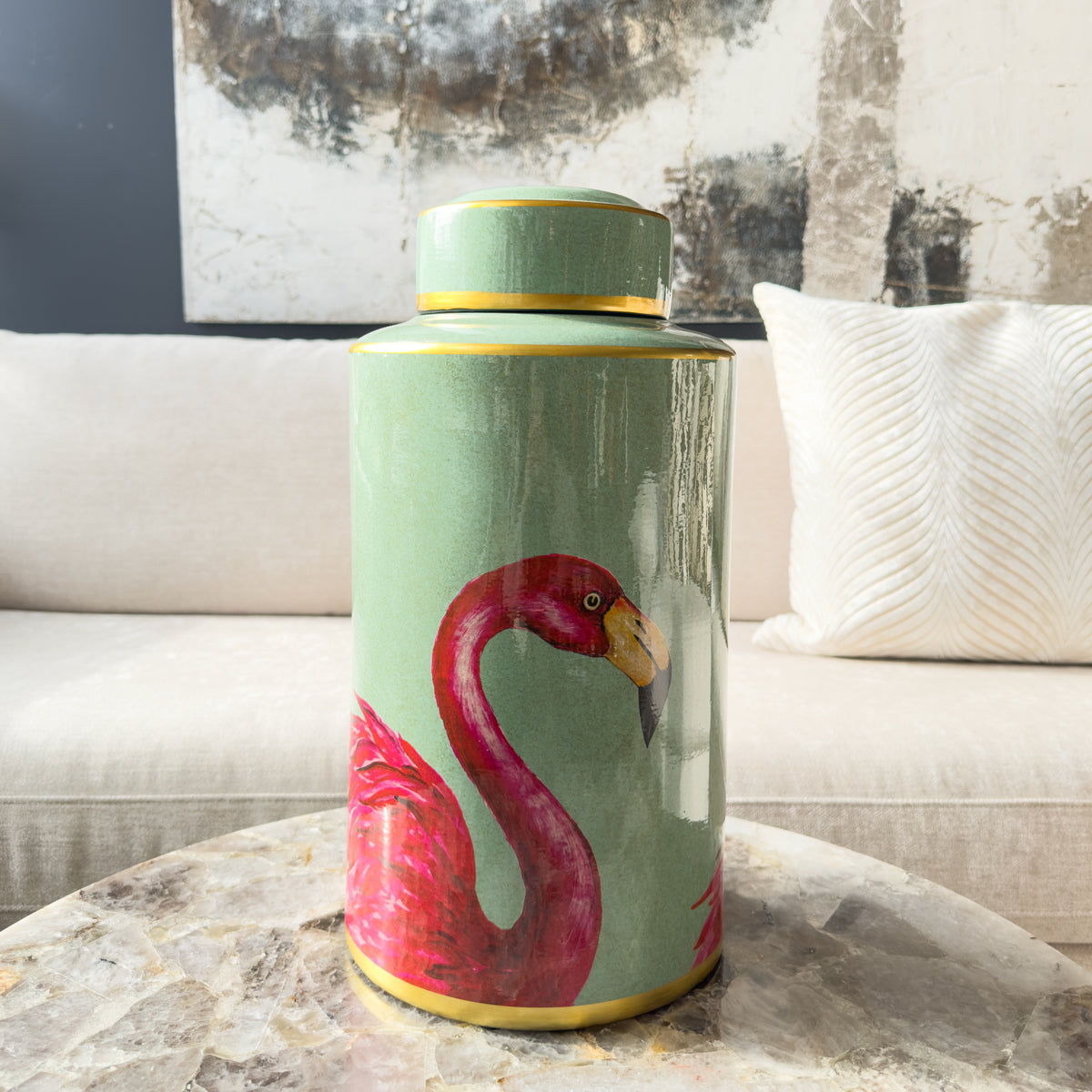 A mint green ginger jar with a vibrant pink flamingo motif, sitting on a quartz coffee table infront of a neutral sofa.