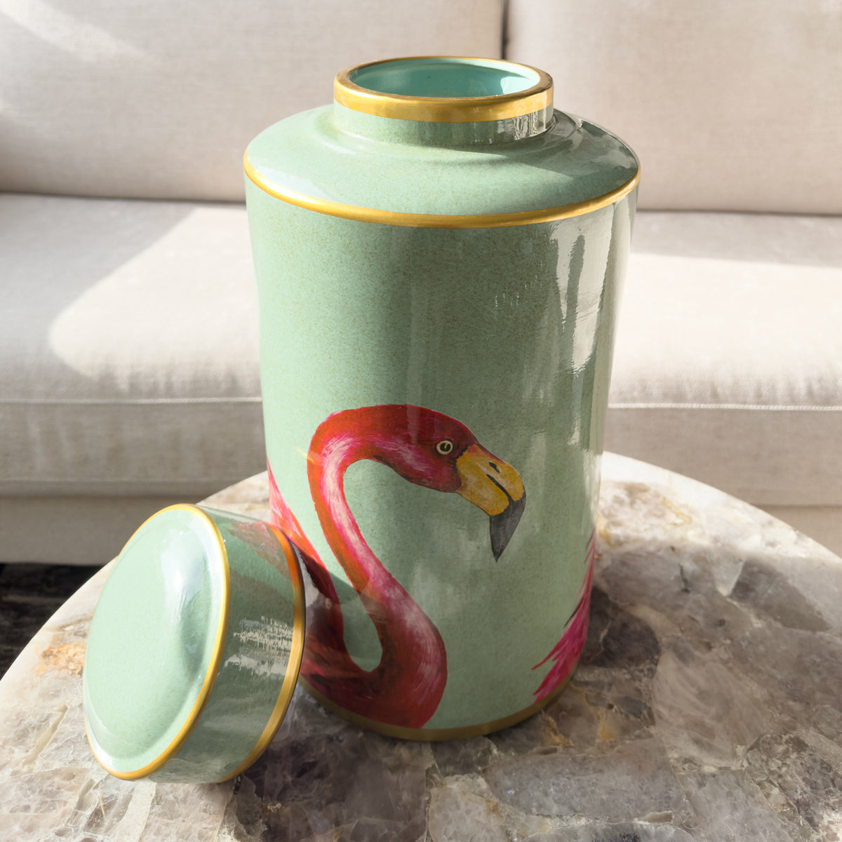 A green ceramic ginger jar with a bright pink flamingo pattern, sitting on a quartz table infront of a cream sofa.