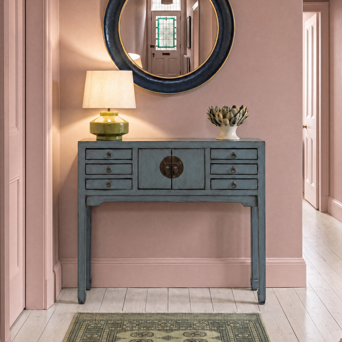 Elegant entryway with a distressed blue console, glowing table lamp, dried floral vase, and round mirror on a dusty pink painted wall.