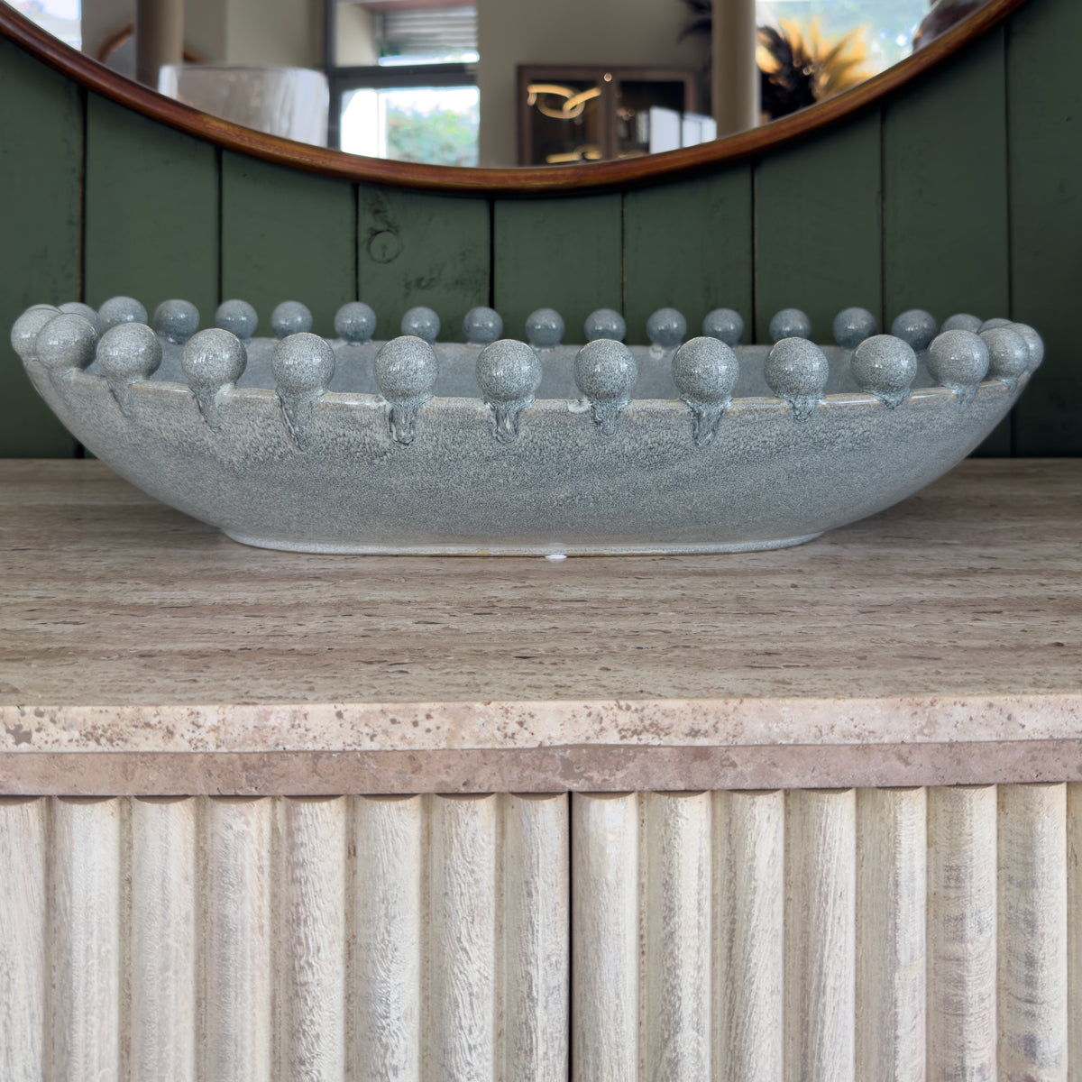 A textured grey blue stone bowl with a ring of rounded knobs sits on a pale stone counter before a curved green-paneled mirror.