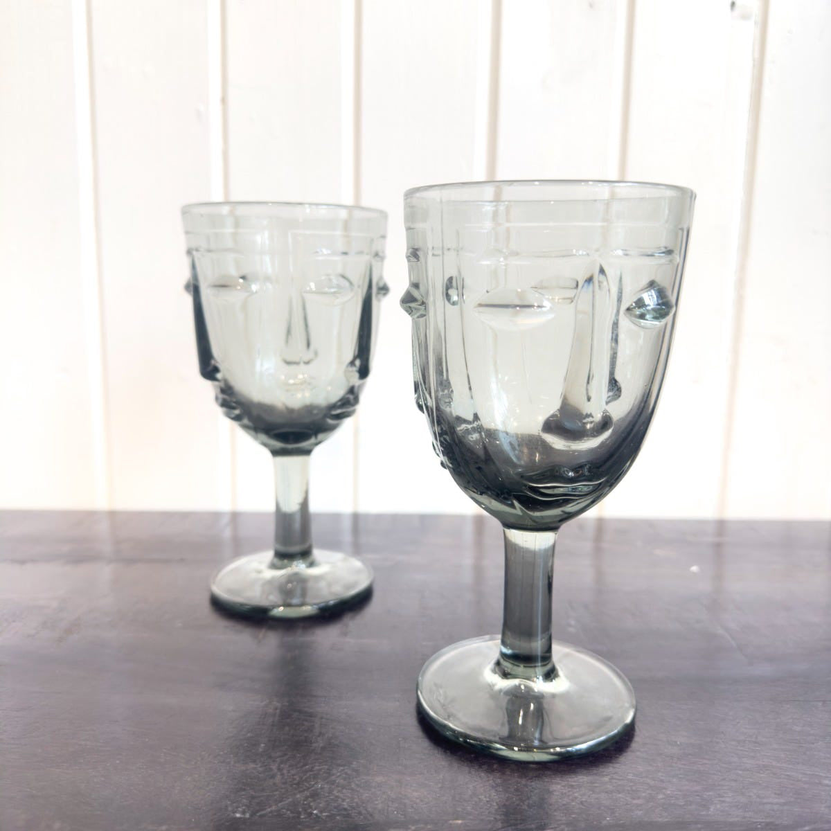 Two grey wine glasses with face motifs standing on a wooden surface against a white background.