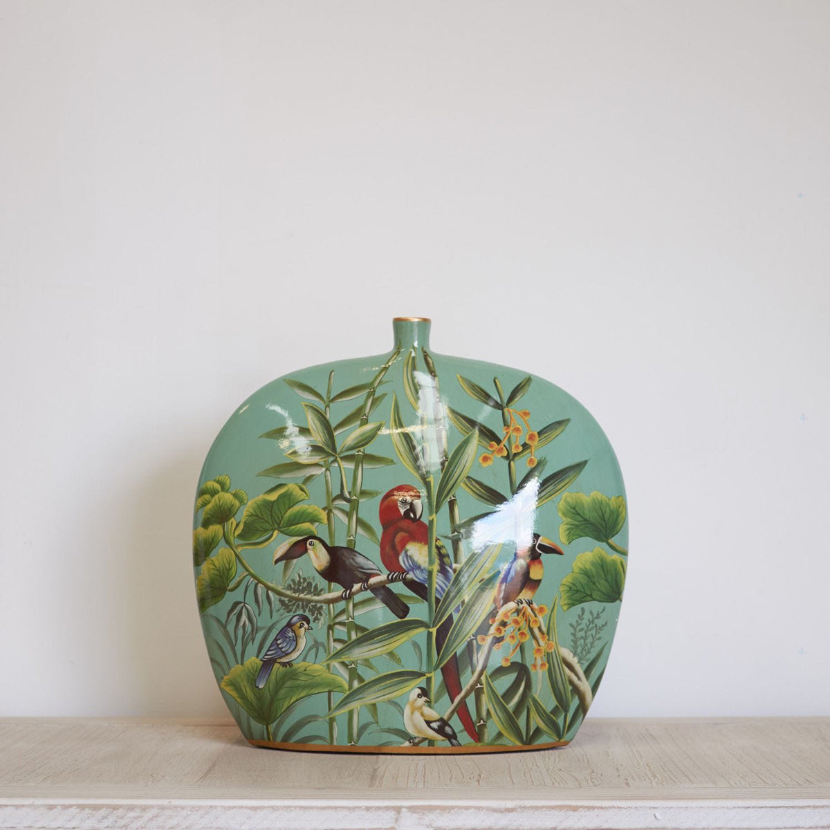 A green bottle vase with a tropical jungle pattern including toucans and a red parrot, sitting on a wooden table.