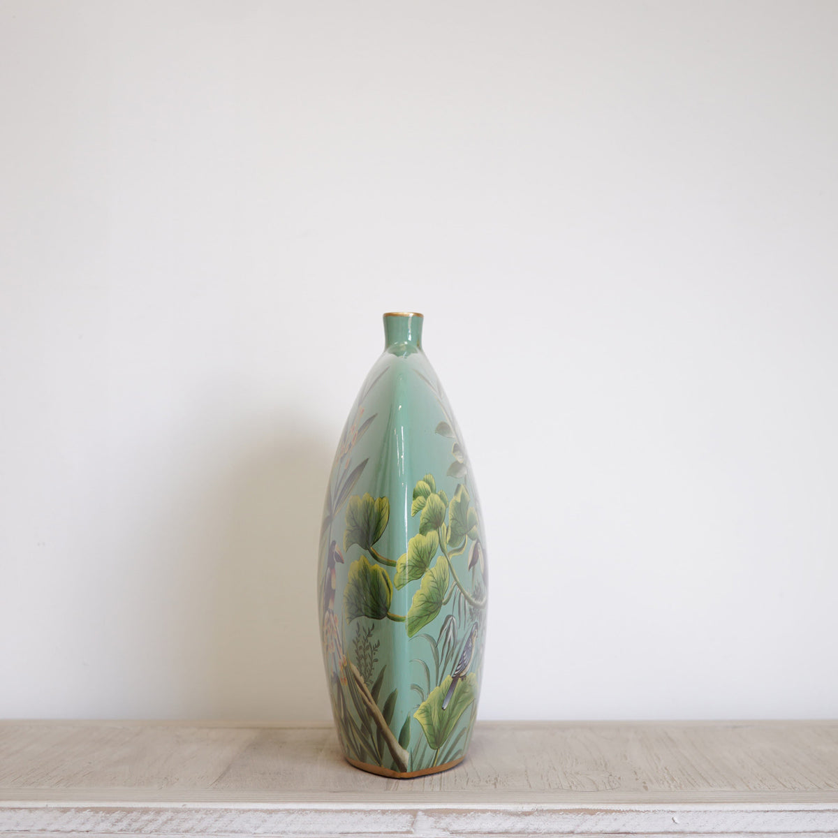 A side view of a green bottle vase featuring a jungle tropical print, sitting on a pale wooden table.