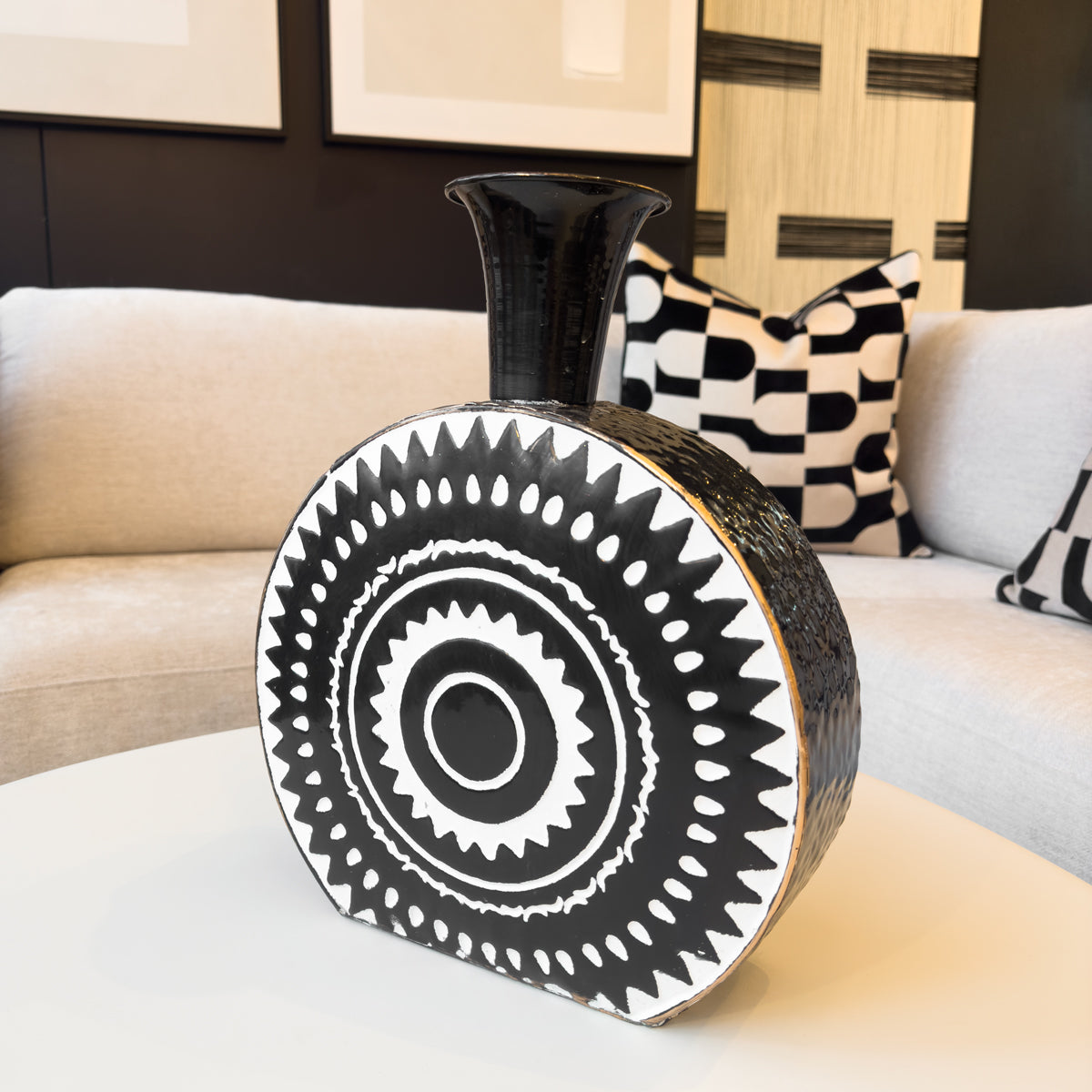 A black and white circular metal vase, featuring a white aztec print, sitting on a table next to a sofa.