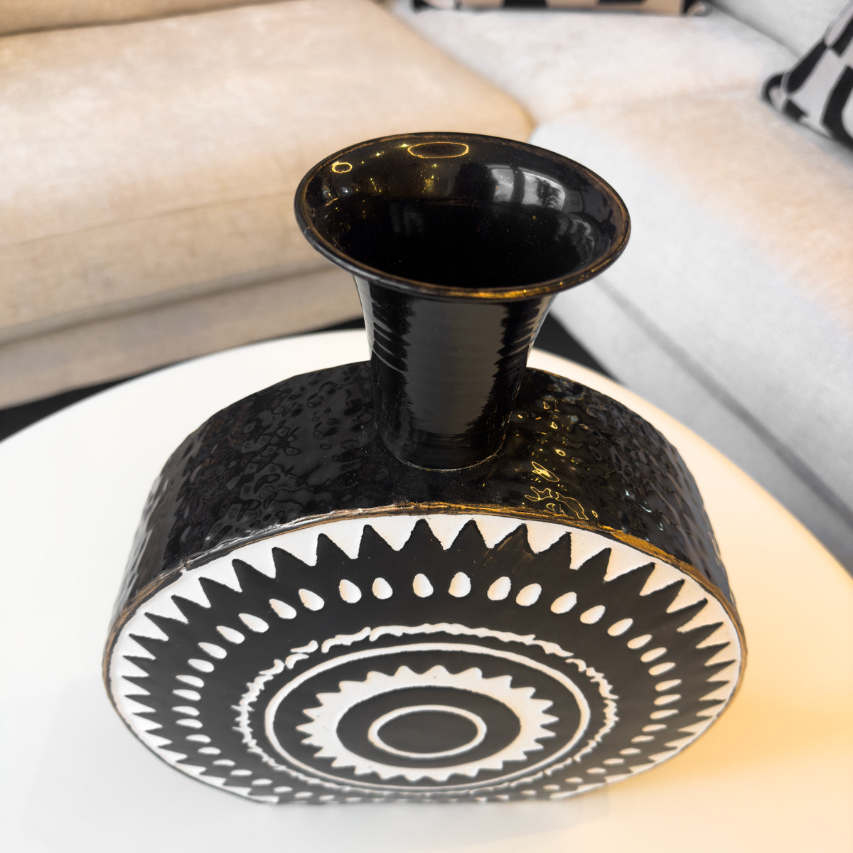 A round monochrome metal bottle vase featuring a white aztec style print, sitting on a table next to a sofa.