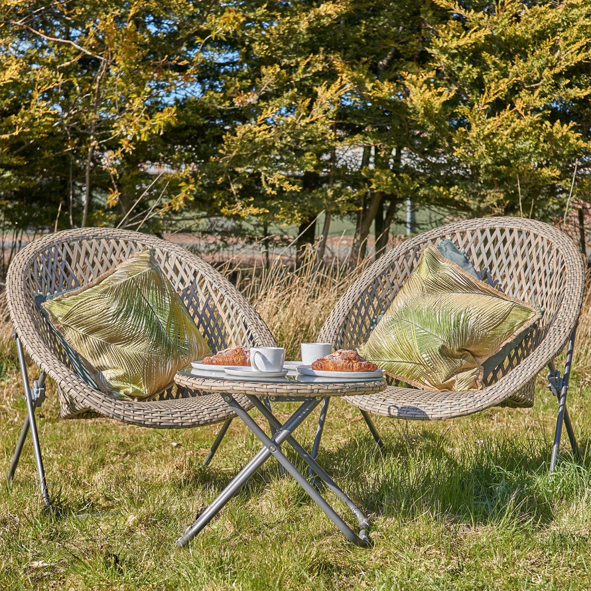 Two rattan chairs with green cushions and a glass top table with a folding mechanism, set on a grassy outdoor area.