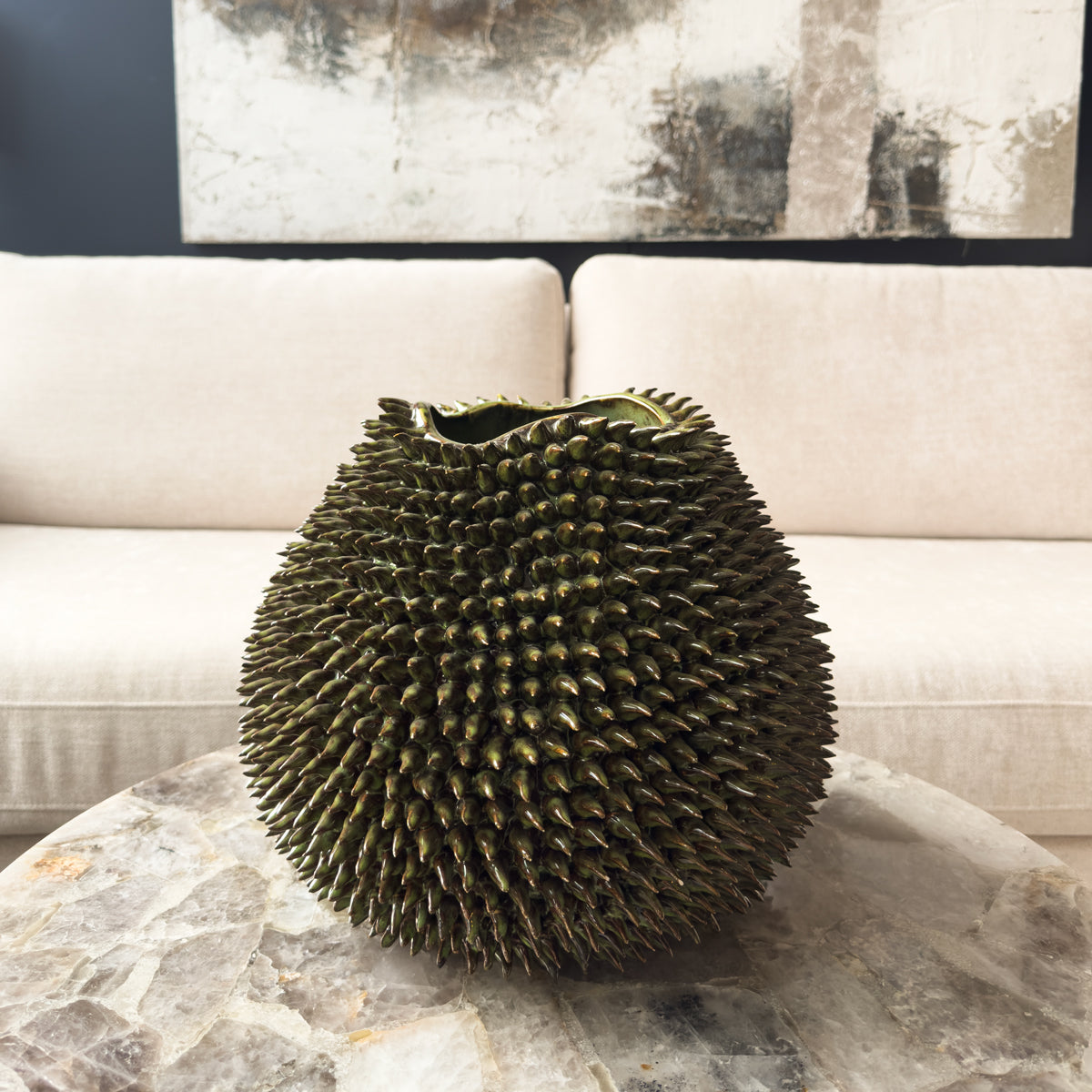 A green sculptural urchin vase sitting on a quartz table infront of a cream sofa and an abstract wall canvas.