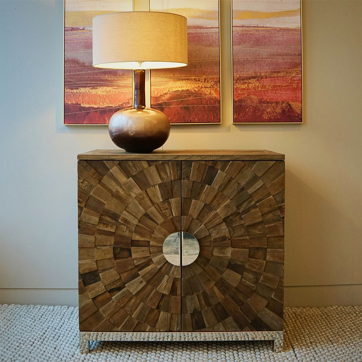 A reclaimed elm and metal 2-door cabinet with spiral geometric carvings, stainless steel legs, and a lamp on top, against a wall with a framed painting.