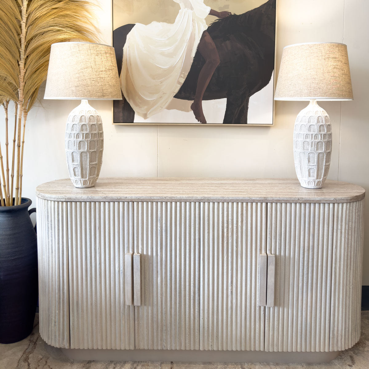 A stylish sideboard with a textured beige finish, and marble top is positioned against a wall. On top, two white lamps with textured bases are flanking a decorative sculpture and a glass candle holder. A painting featuring a woman in a white dress hangs above.