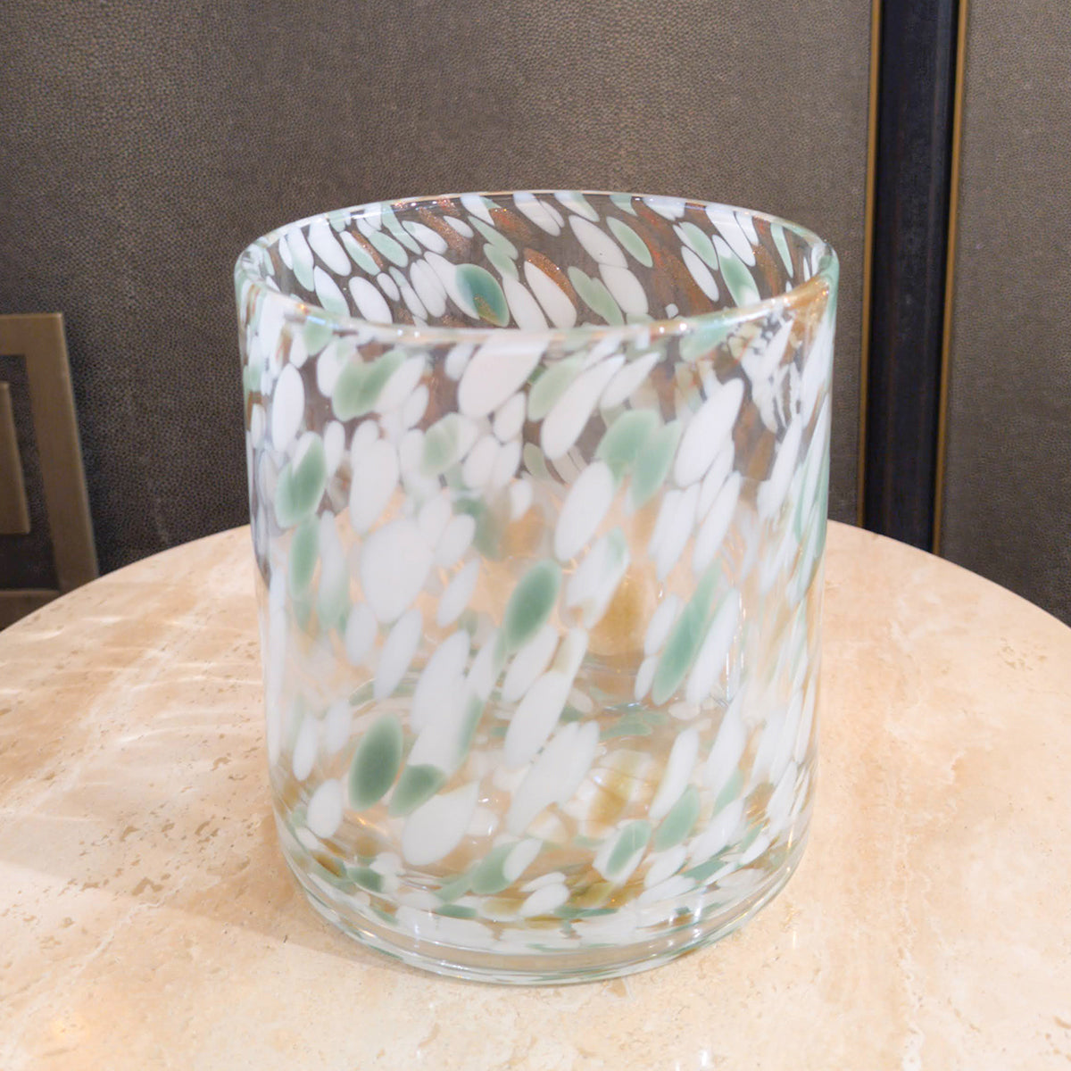 A cylindrical glass hurricane with a confetti pattern in shades of sage green, white, and metallic gold.