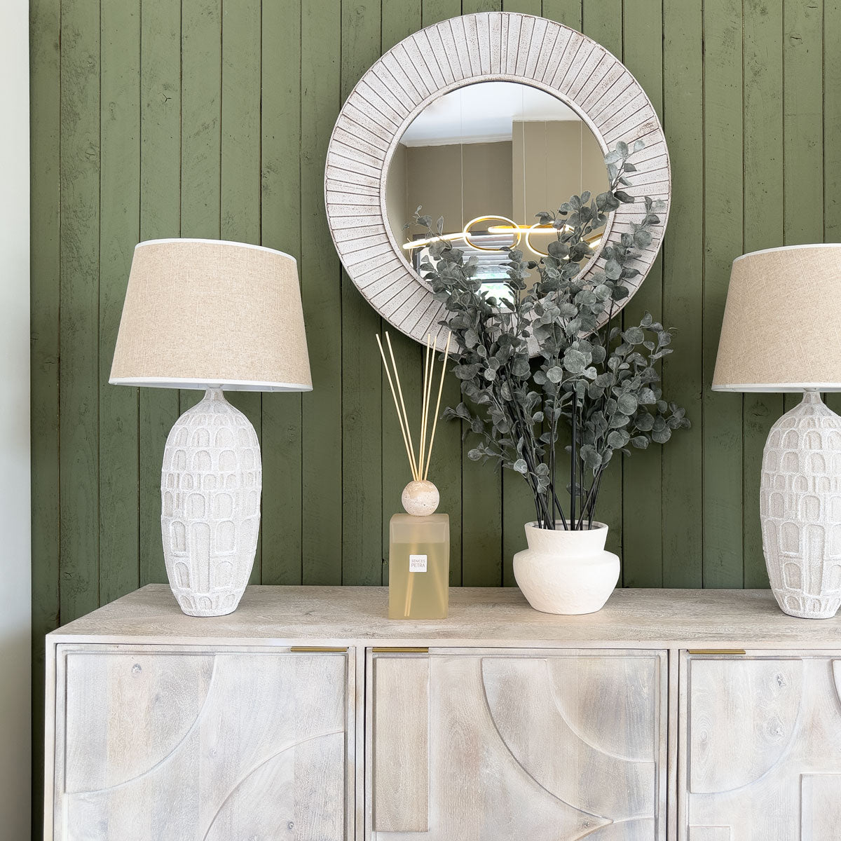 A light wooden sideboard with three doors featuring a geometric design, placed against a green panelled wall. On top, there are two white lamps, a glass diffuser and dried foliage in a white planter.