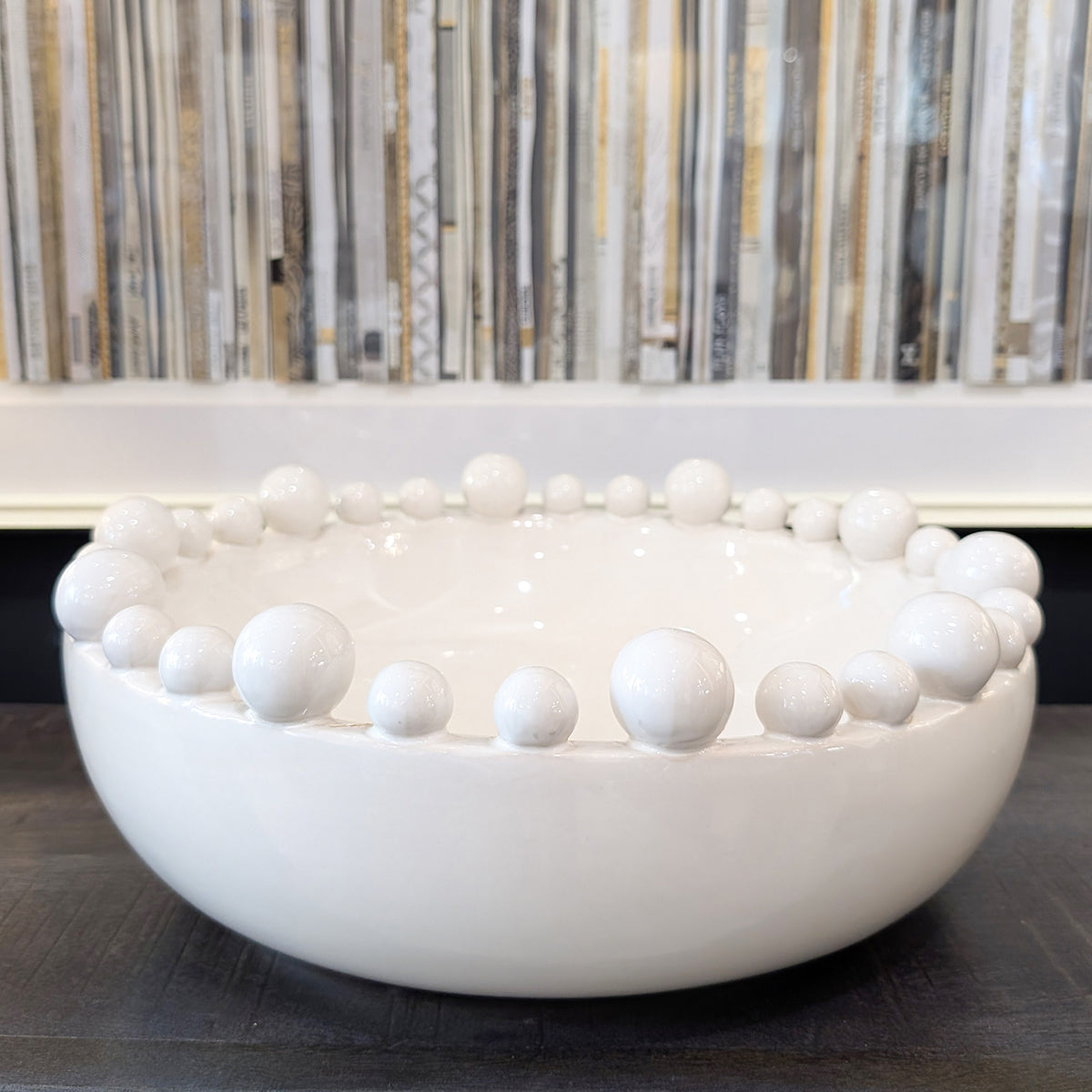 A white bobble design bowl sitting on a dark wood tabletop in front of a book themed artwork.