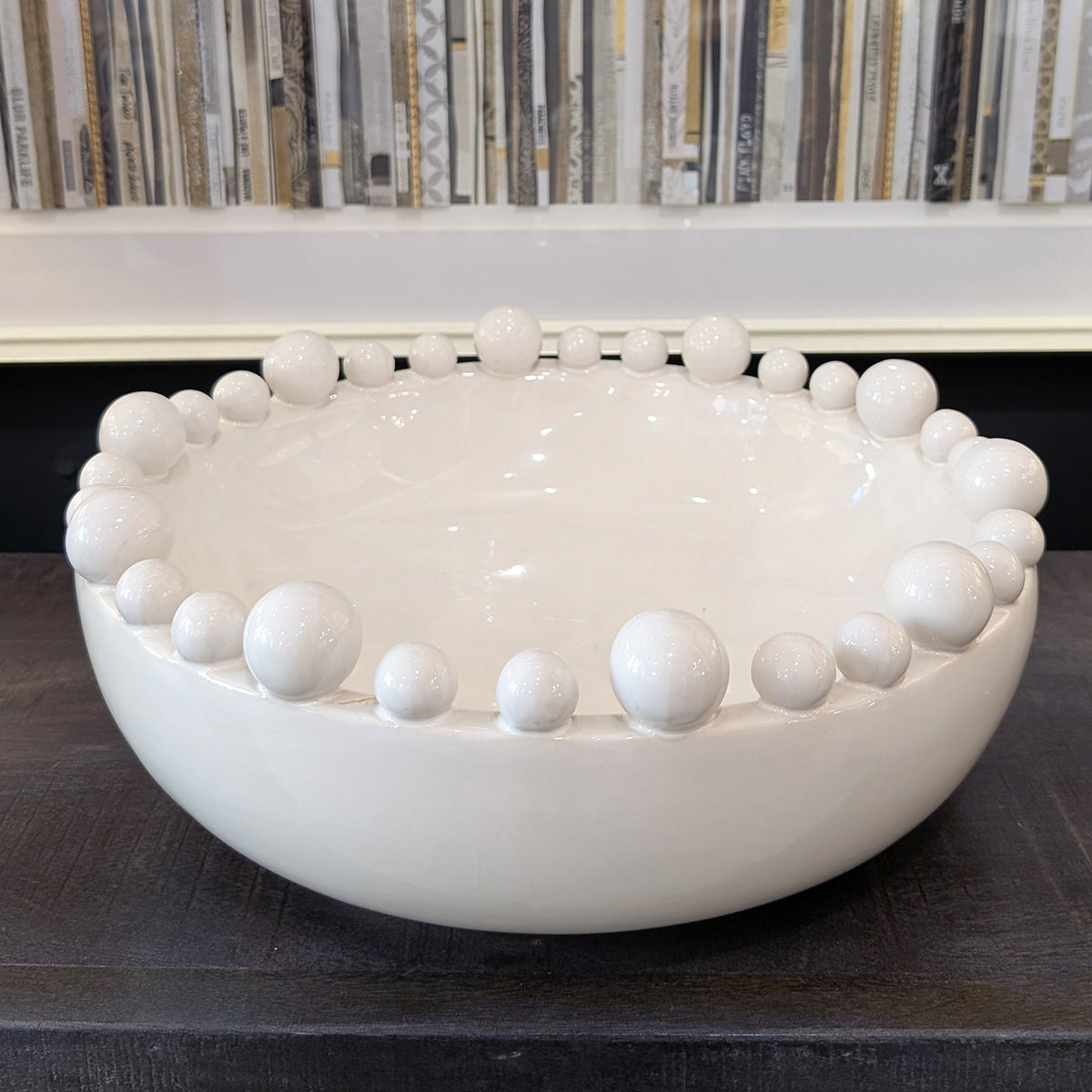 A close up of a white bowl with bobble details around the rim, sitting on a black console table beside a bookshelf picture.