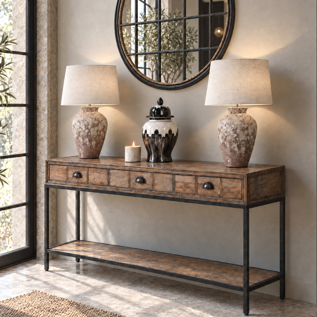 A rustic console table with a distressed brown wood finish and black metal frame, styled with two matching table lamps, a small ceramic jar, and a candle in a home entryway setting.