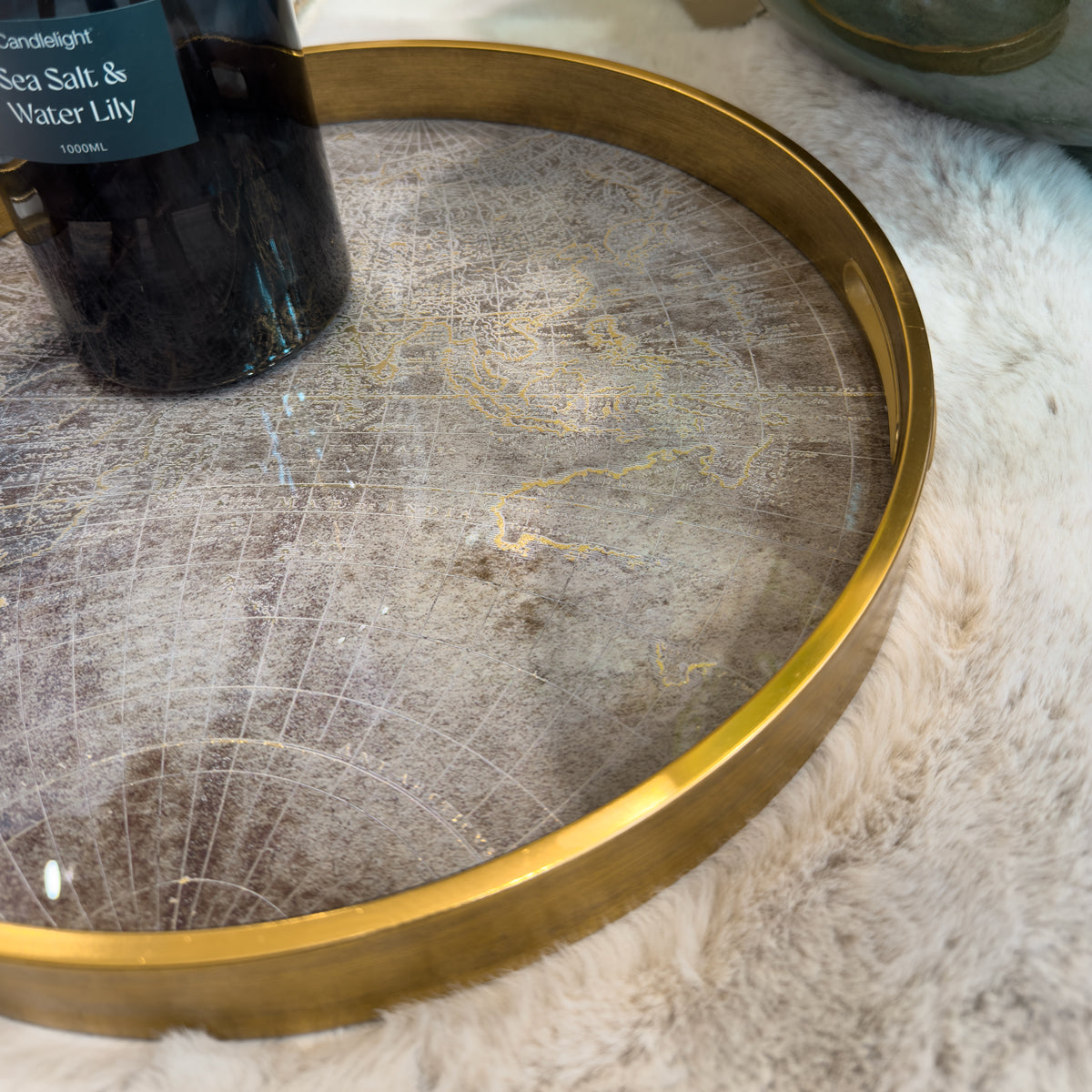 Zoomed in image of the gold round atlas tray showing the map design, sitting on a cream fluffy rug. To the left-hand side there is the bottom of a reed diffuser.