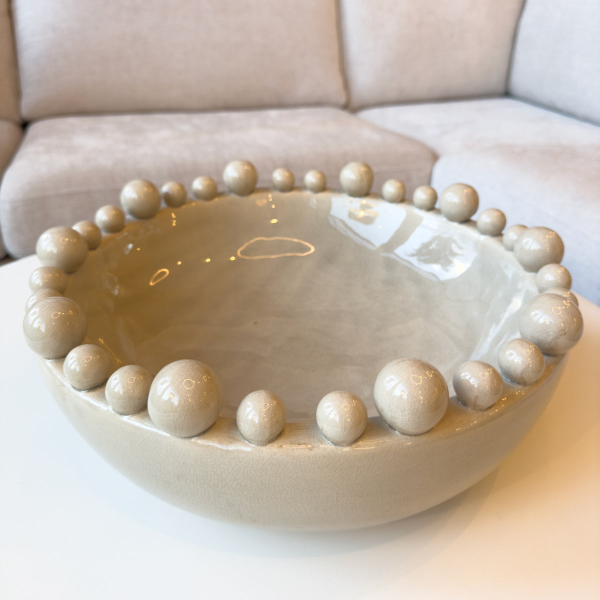 A cream gloss bowl with bobbles around the rim, sitting infront of a cream coloured corner sofa.