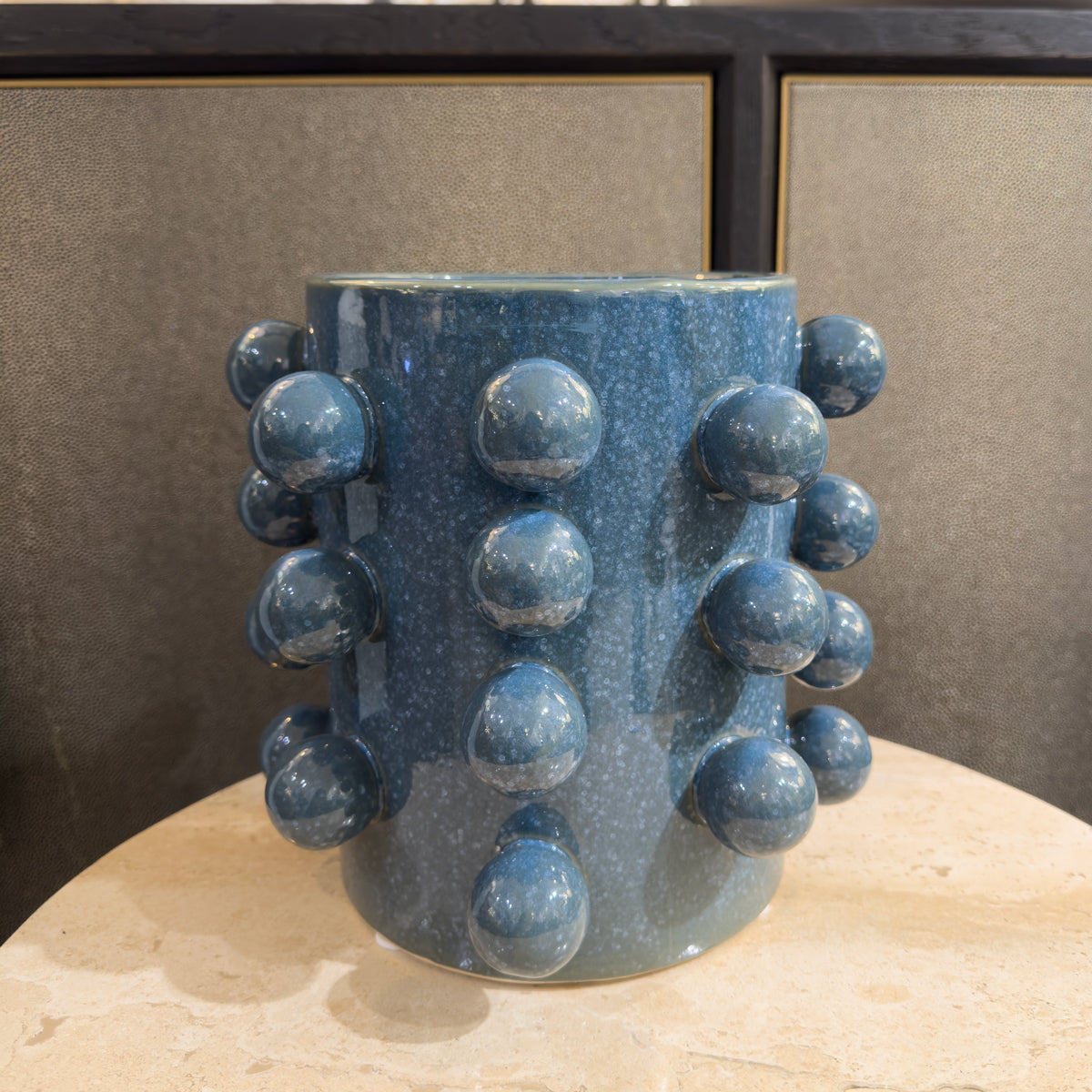 A blue plant pot with ceramic ball embellishments, sitting infront of a beige & black sideboard.