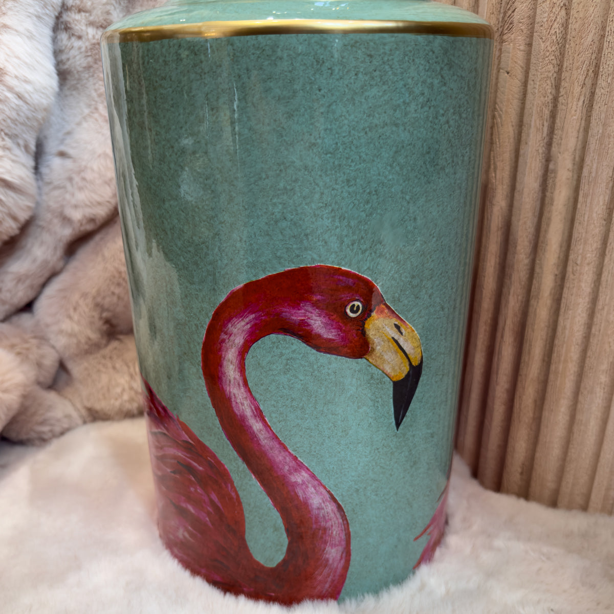 A cylindrical ceramic jar featuring a painted hot pink flamingo against a light teal background. The flamingo's head and neck are prominently displayed, with a small portion of the body visible. The vase is placed on a soft surface.