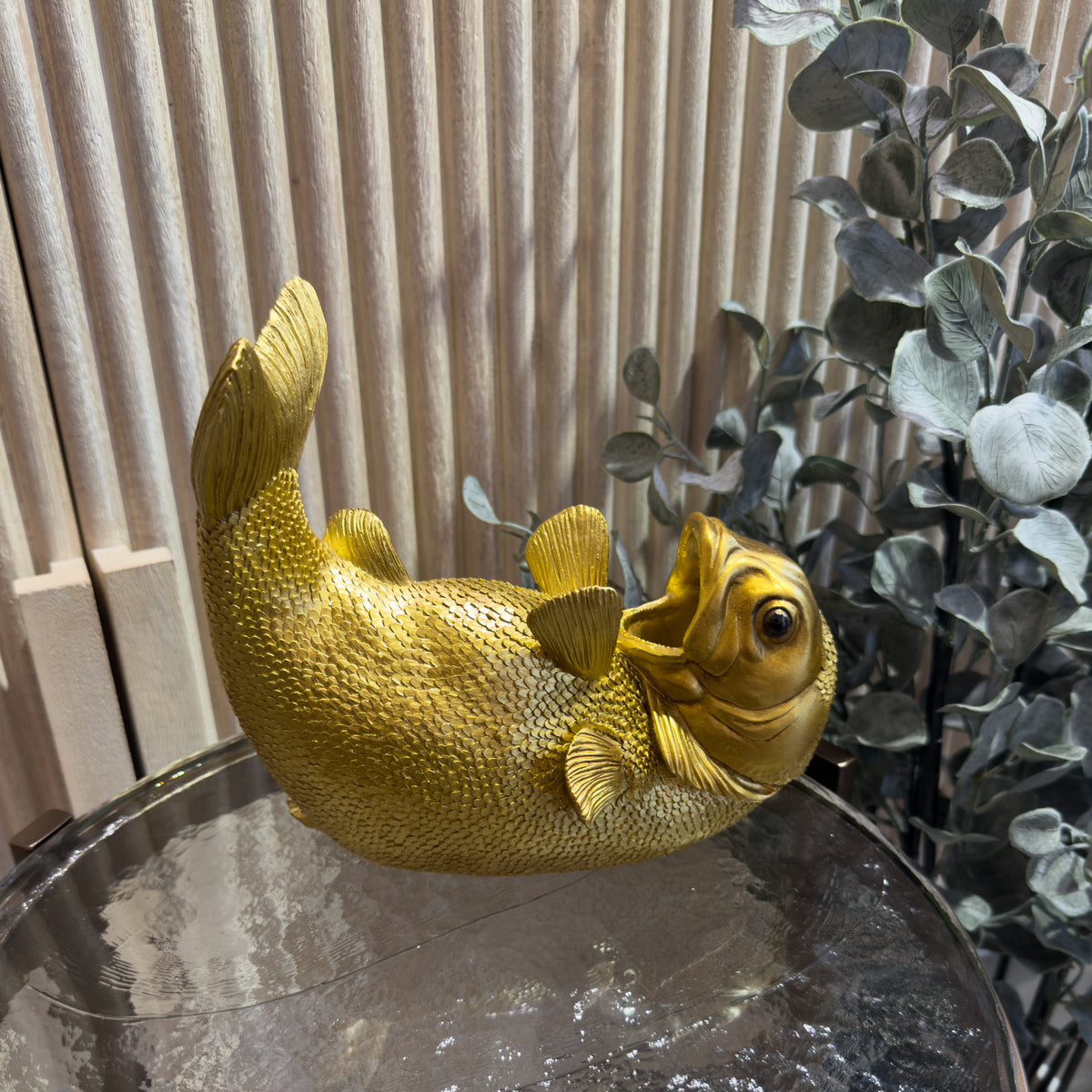 Golden fish wine bottle holder with raised tail rests on a glass table, surrounded by leafy plants and wood paneling, creating a whimsical indoor scene.