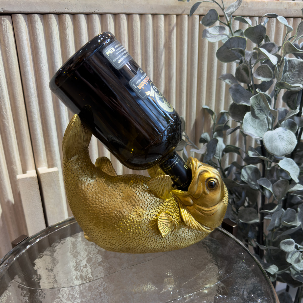 Whimsical golden fish wine bottle holder balances a dark bottle on its back, displayed on a glass table beside leafy plants in a cozy setting.
