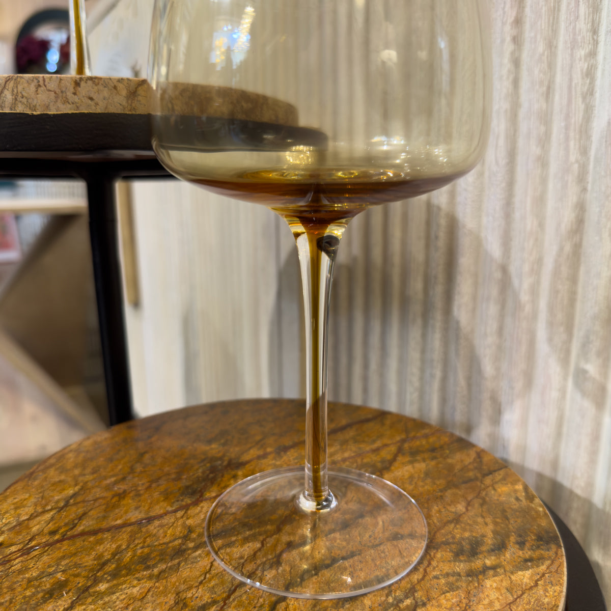 One glass wine glass with golden flooded detail stand on a round brown marble table beside a washed wood sideboard.