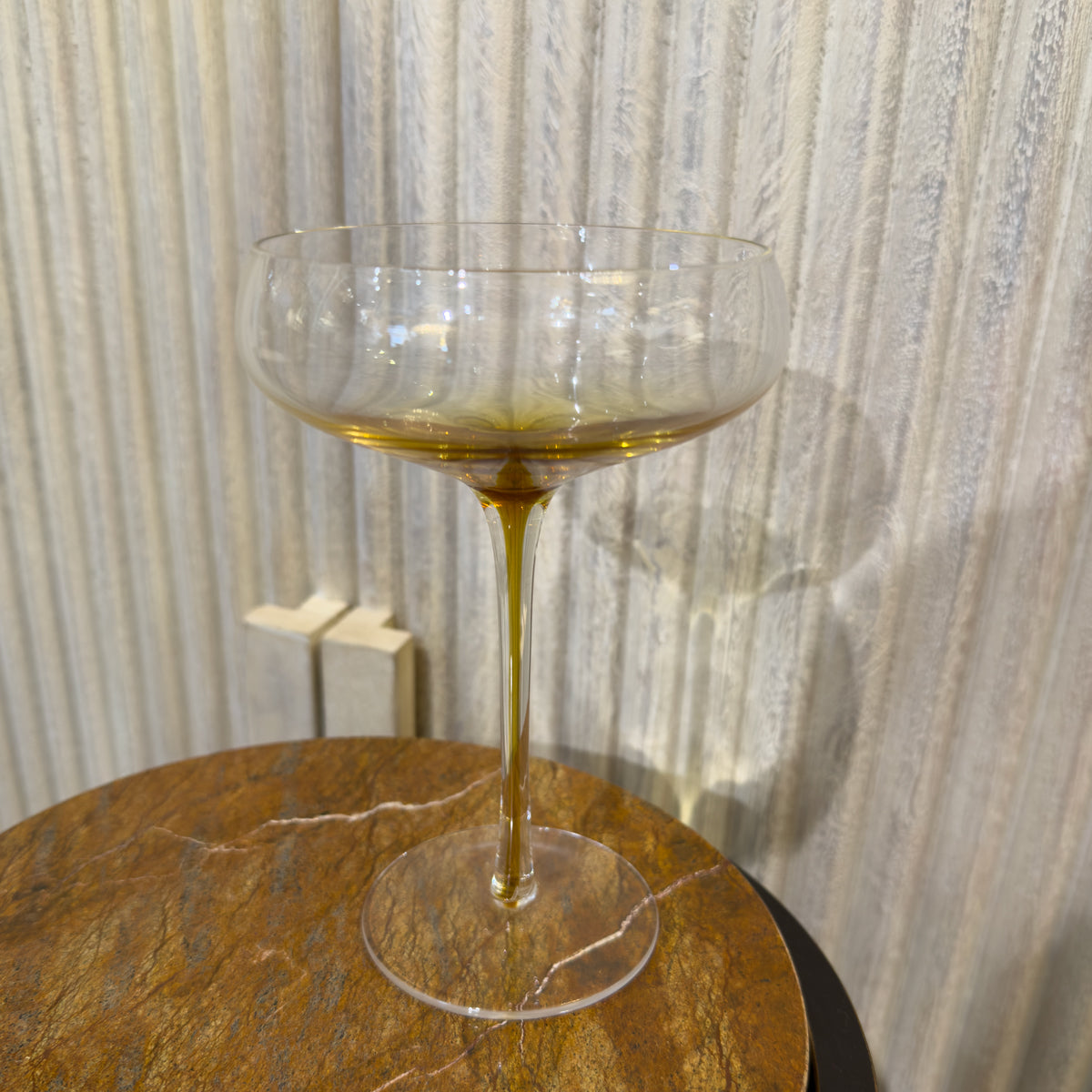One glass clear cocktail glasses with golden stem stand on a round marbled table against pale textured sideboard.