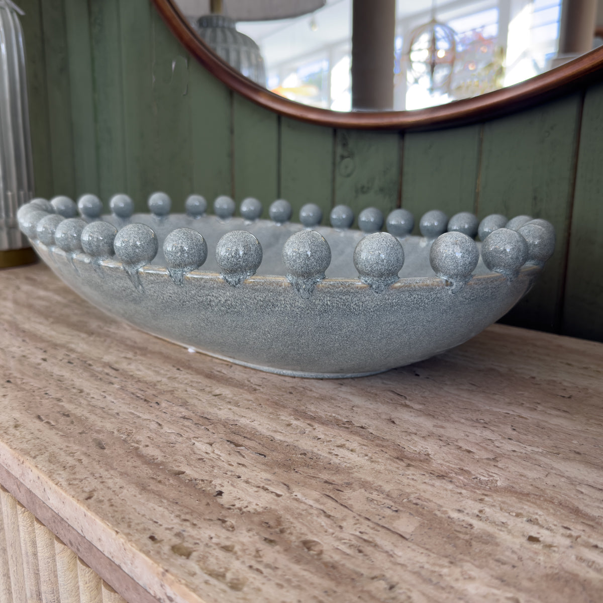 A textured grey blue stone bowl with a ring of rounded bobbled edges sits on a pale stone counter before a curved green-paneled mirror, creating a calm, refined bathroom scene.