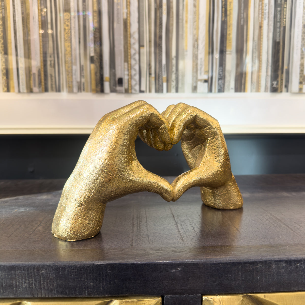 Gold sculpture of two hands forming a heart shape on a dark surface with a patterned picture in the background.