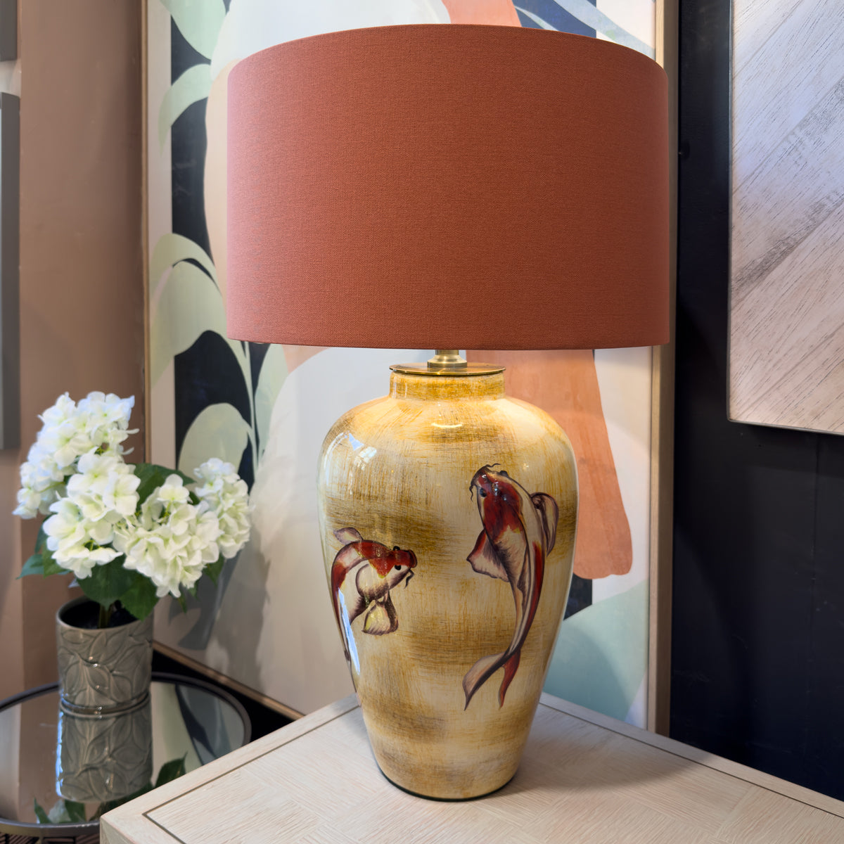 A table lamp with a glossy beige ceramic base decorated with two koi fish, topped with a large terracotta-colored drum shade, shown on a light wood console table next to a vase of white flowers.