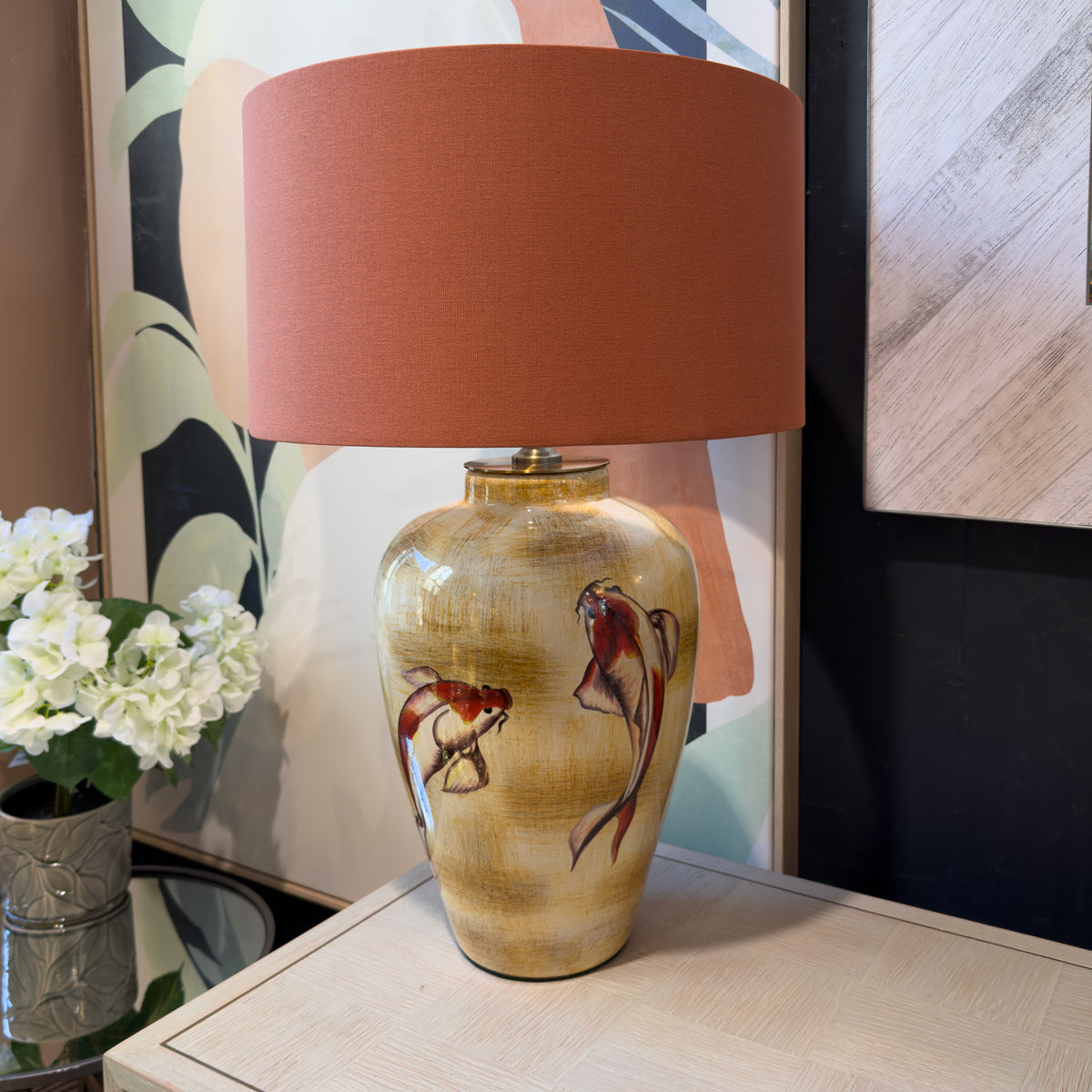 A table lamp with a warm terracotta drum shade and a glossy beige ceramic base decorated with painted koi fish, shown on a light wood console table in a styled interior setting.