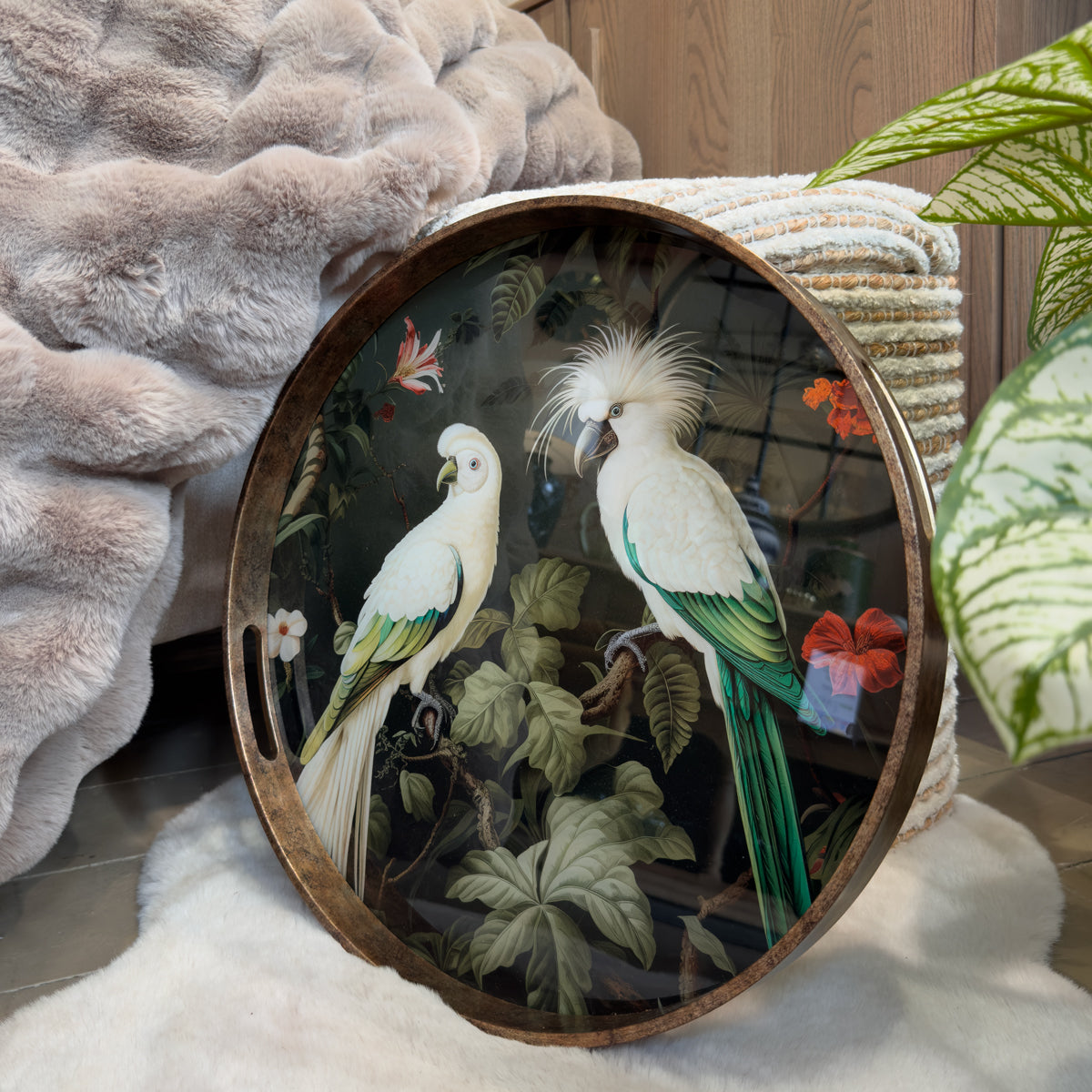 Round decorative tray featuring two elegant white cockatoos with soft green and turquoise feather detailing, perched amongst lush tropical leaves and vibrant botanical flowers. Set against a rich black background with exotic foliage, butterflies and colourful blooms, the artwork is framed by a warm bronze-effect rim. Displayed on a soft faux fur throw beside a wood-panel wall, potted green plant, this statement tray brings luxurious botanical style and tropical elegance to any contemporary home.